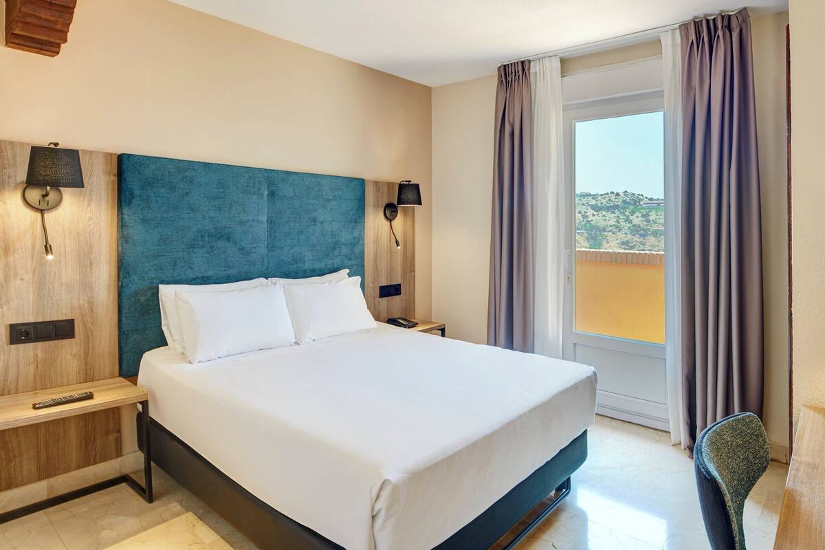 Une chambre d’hôtel avec un lit, deux tables de chevet, une lampe, une chaise et une vue sur l’extérieur.