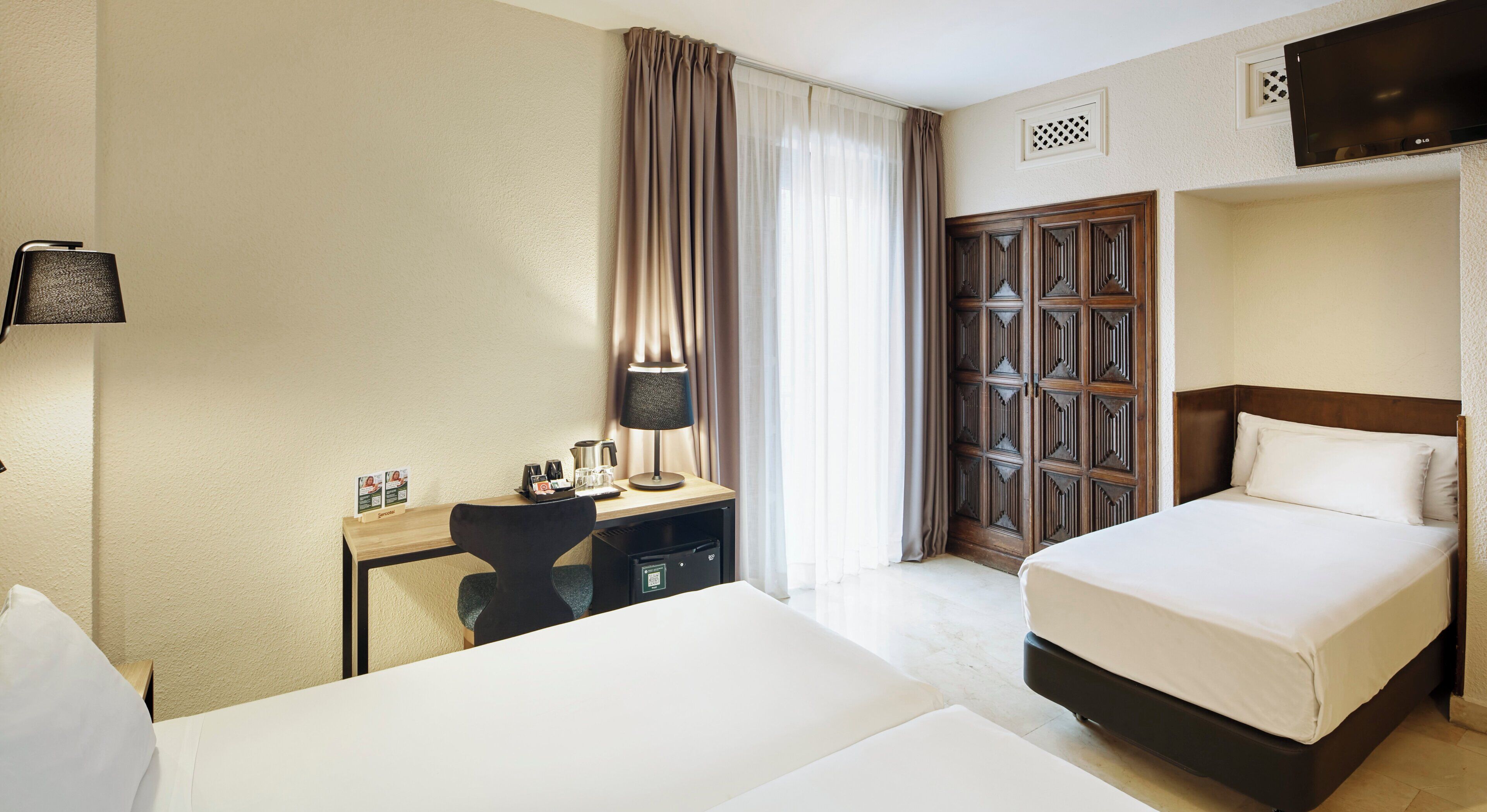 Habitación de hotel con cama, escritorio con lámpara, silla, armario y televisión en la pared.