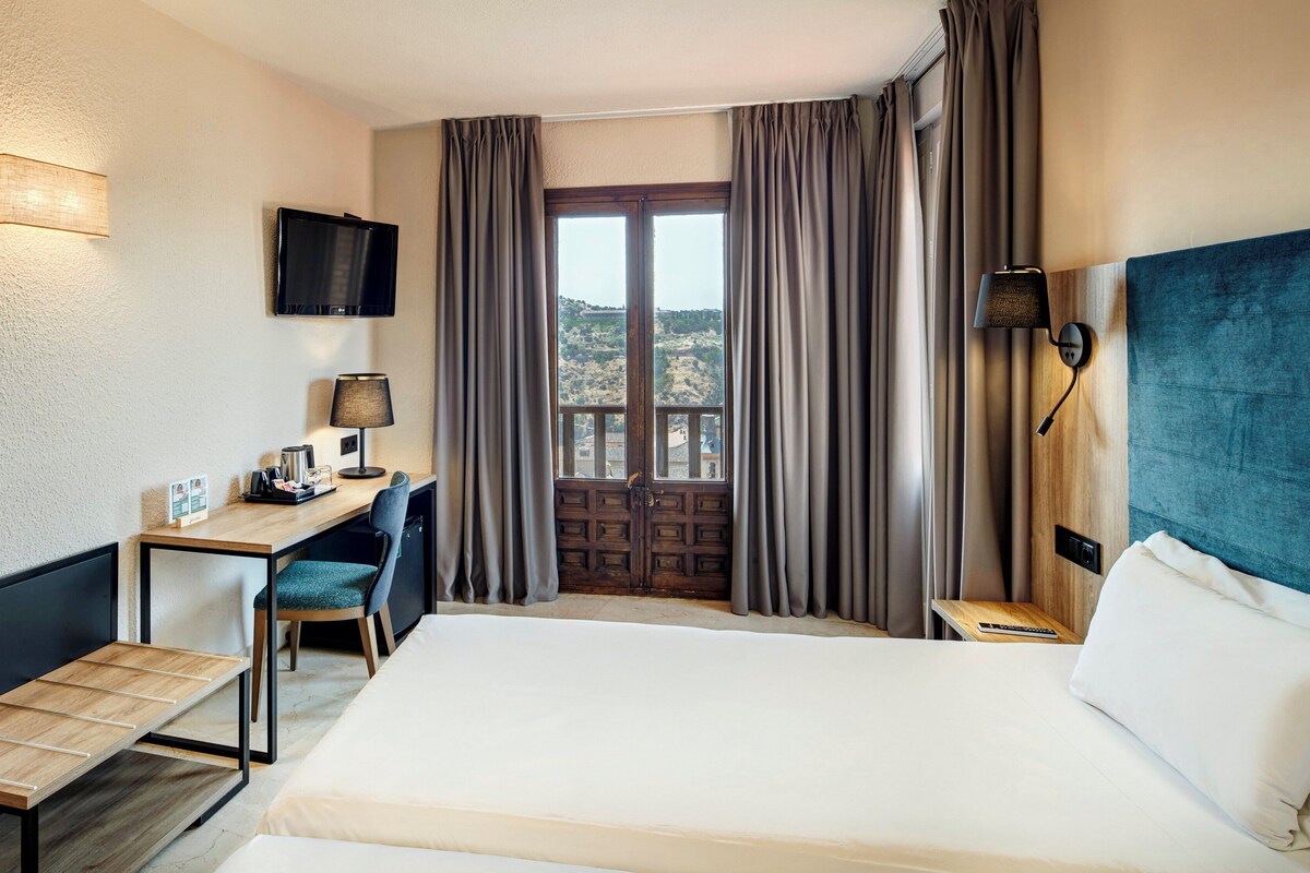 A hotel room with a bed, desk, chair, lamp, and a TV on the wall.