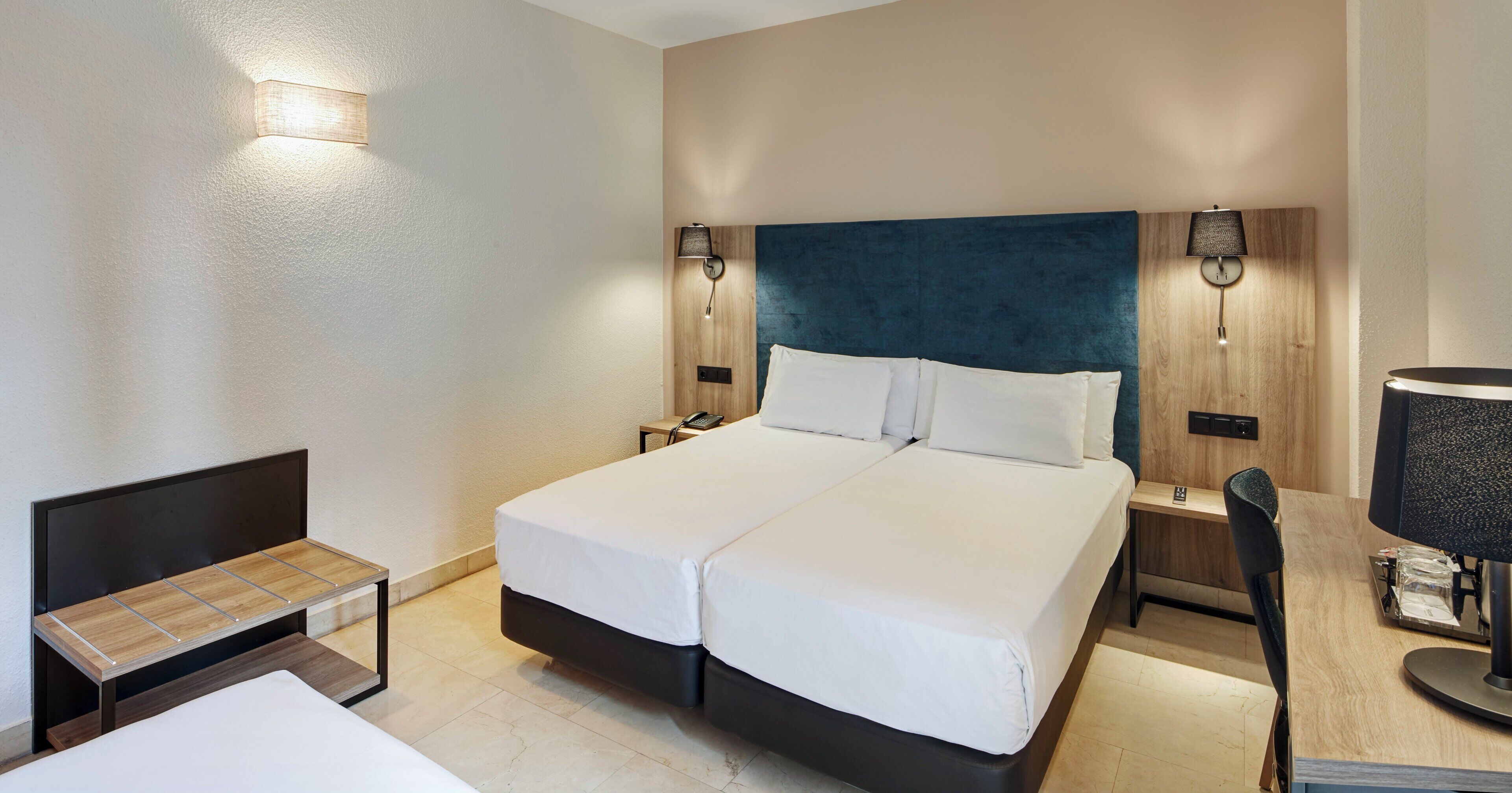 Habitación de hotel con cama doble, mesitas de noche, escritorio con silla y dos lámparas de pared.