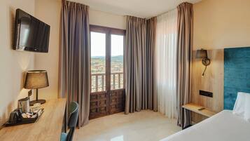 Una habitación de hotel con una cama, un escritorio con lámpara, televisión y vistas a un paisaje urbano a través de una ventana con cortinas.