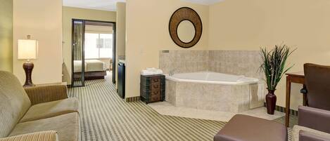 A hotel room with a large bed, a sitting area with a sofa and armchairs, a bathroom with a bathtub, and a mirror on the wall.