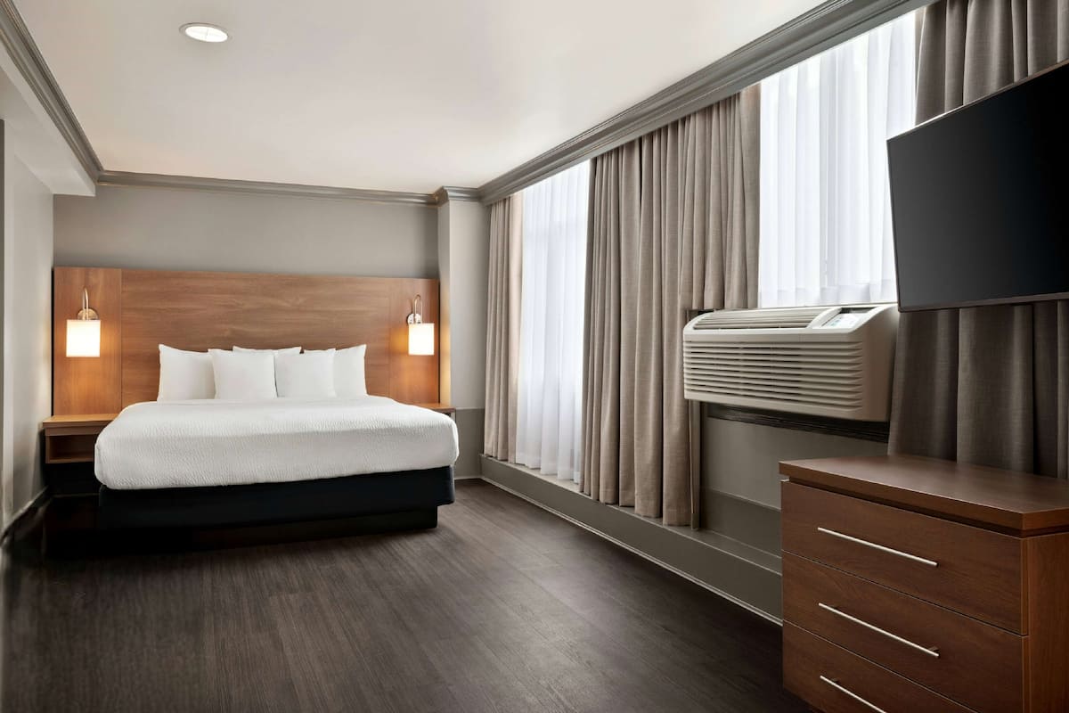 A modern hotel room with a large bed, bedside lamps, a flat-screen TV, a dresser, and a large window with curtains.