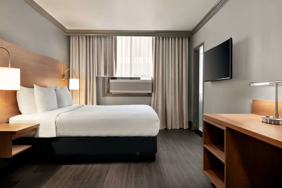 A hotel room with a bed, bedside tables, a desk, a TV, an air conditioning unit, and a window with curtains.