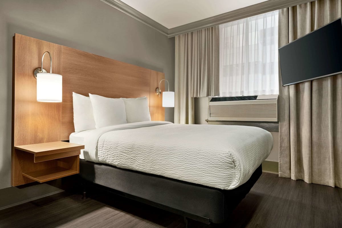 A hotel room with a large bed, two bedside tables, a flat-screen TV, an air conditioning unit, and a window with curtains.