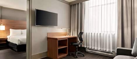 Una habitación de hotel con cama, un escritorio y una silla, televisor y un amplio ventanal con cortinas.