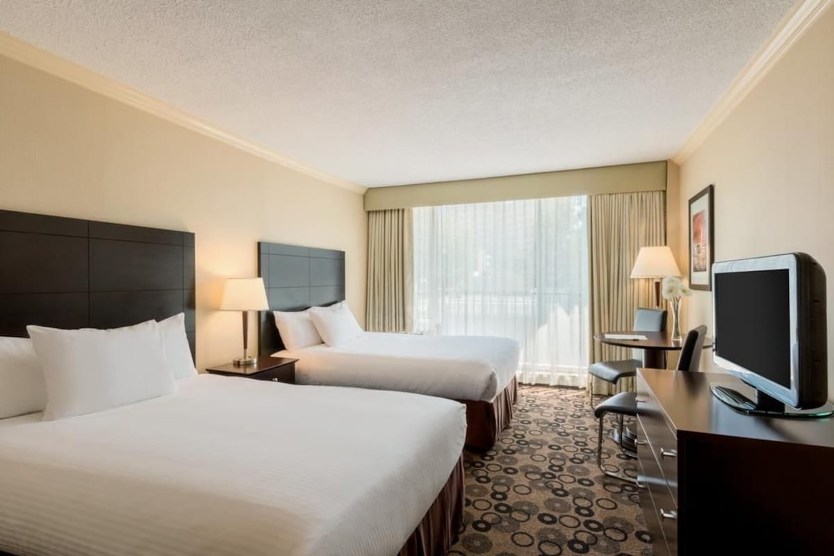 A hotel room with two beds, a desk with a TV, a chair, a lamp, and a large window with curtains.