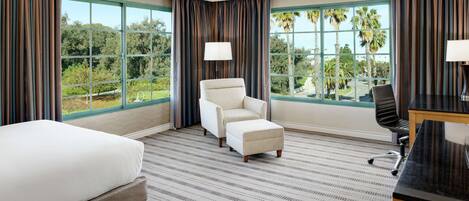 A hotel room with a bed, a chair, a desk, and large windows offering a view of palm trees and greenery.