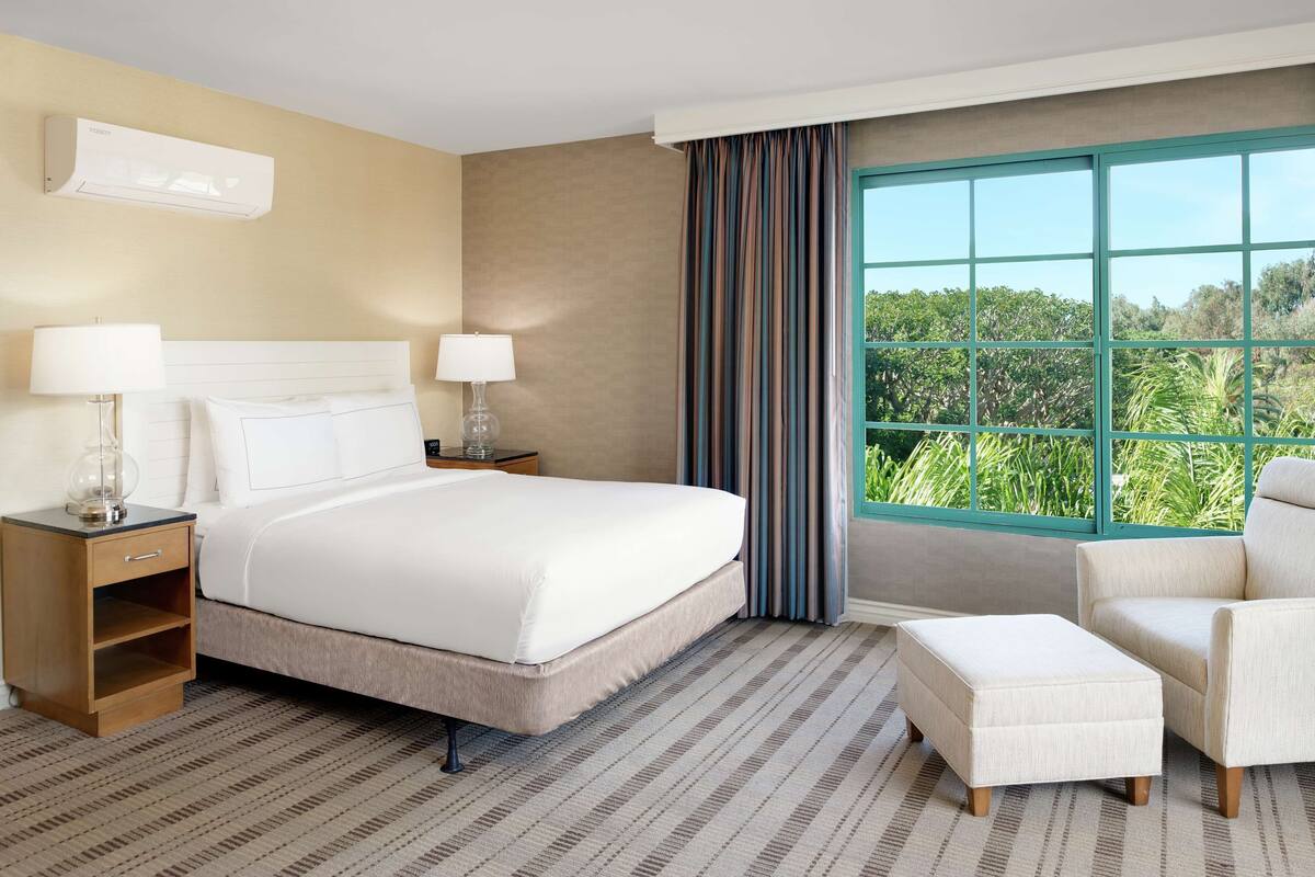 Une chambre d’hôtel avec un grand lit, deux tables de chevet avec des lampes, une chaise, un pouf et une fenêtre donnant sur un paysage verdoyant.
