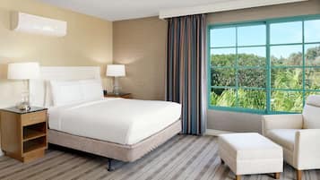 Una habitación de hotel con una cama grande, dos mesitas de noche con lámparas, una silla, un reposapiés y una ventana con vistas a zonas verdes.