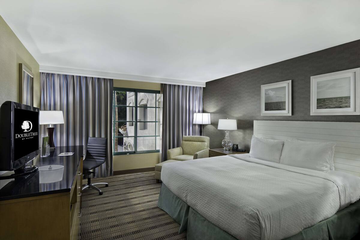 Une chambre d’hôtel comprenant un lit, un bureau avec un ordinateur, deux chaises, une télévision et une fenêtre avec des rideaux.