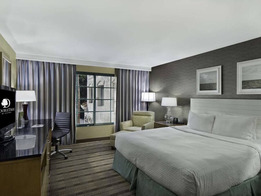 A hotel room with a bed, a desk with a computer, two chairs, a TV, and a window with curtains.