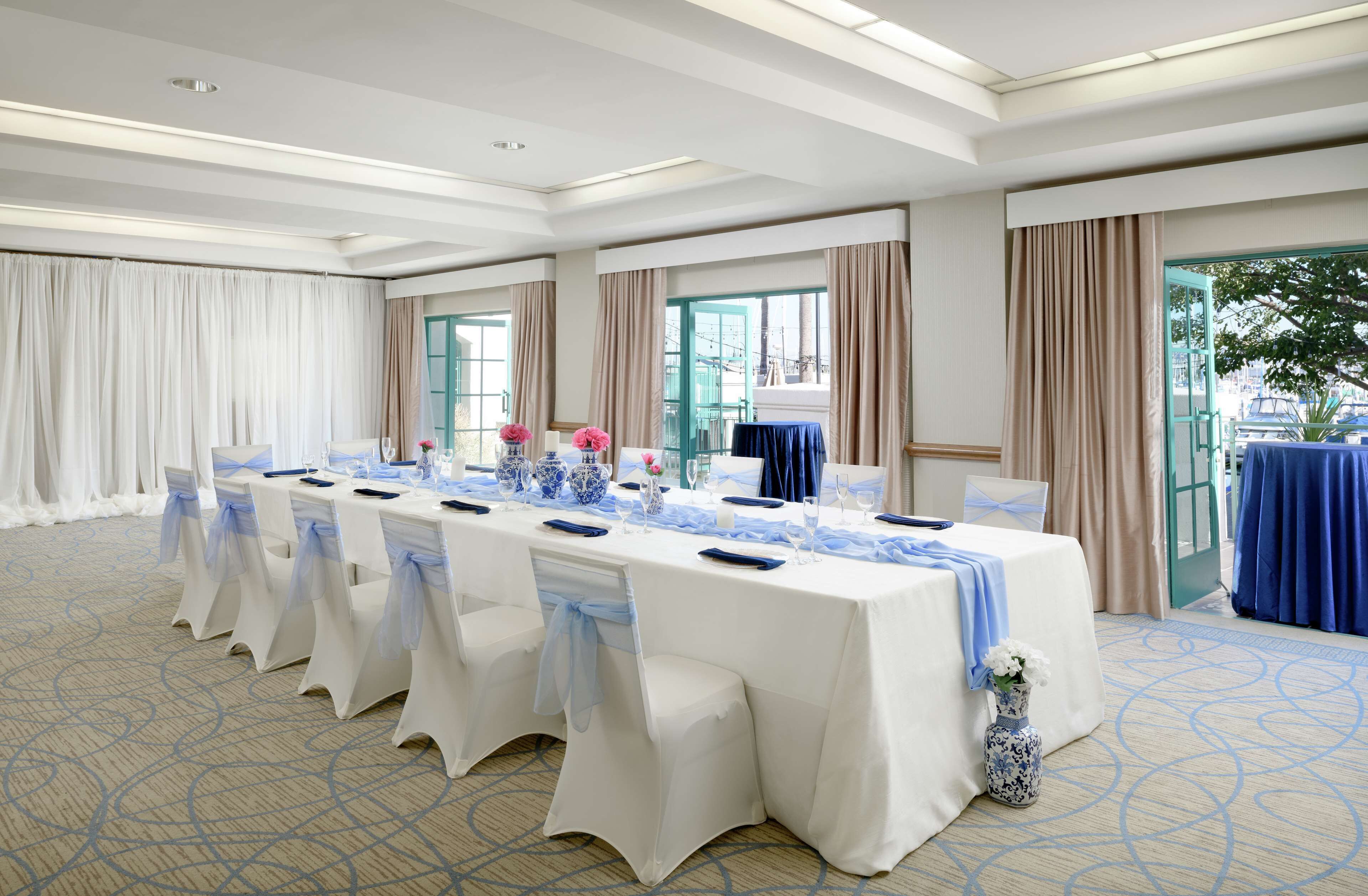 A banquet hall with a long table set for a formal event, featuring white tablecloths, blue sashes, and floral centerpieces.