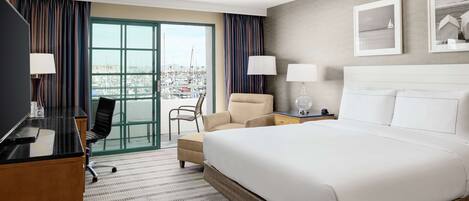 Habitación de hotel con una cama grande, un escritorio con silla, un sofá y vista a un puerto deportivo por la ventana.
