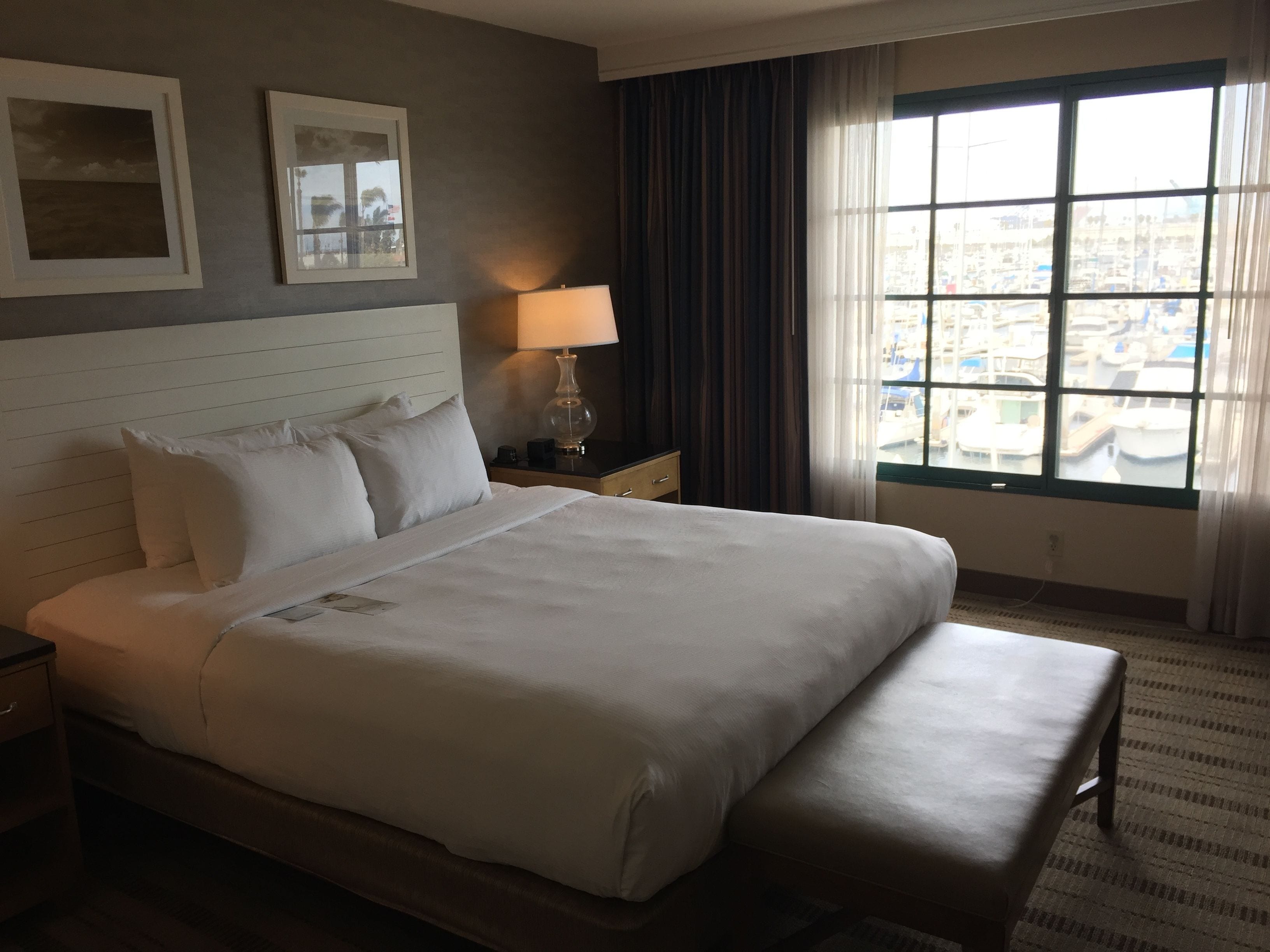 A hotel room with a large bed, a bedside table with a lamp, a window with a view of the city, and two framed pictures on the wall.