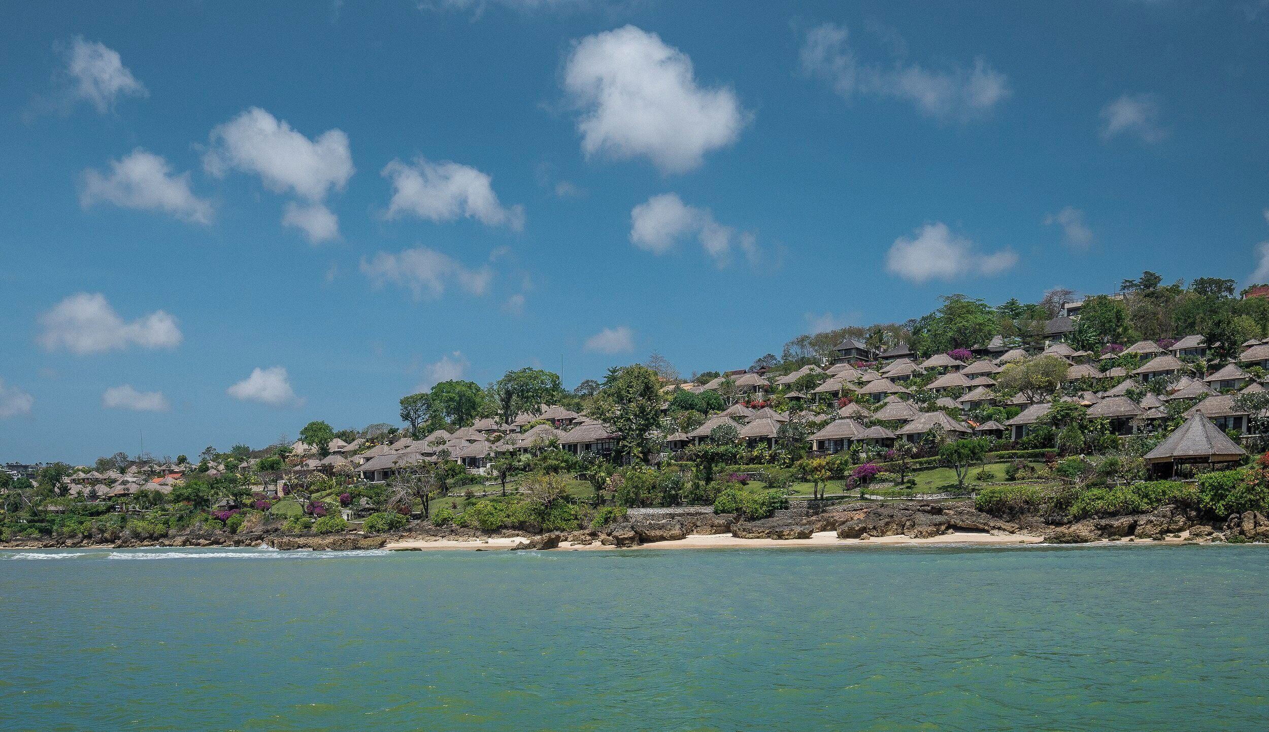 Un complejo turístico costero con bungalows de techo de paja, una playa arenosa y aguas azules cristalinas.