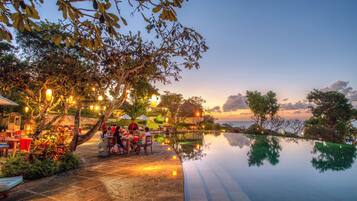 Un área de comedor al aire libre con mesas y sillas, una piscina y árboles con luces.