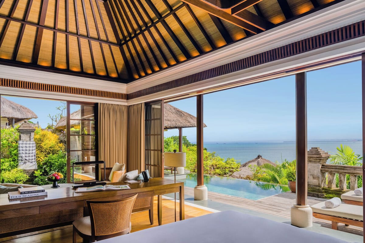 Ein Zimmer mit großem Fenster mit Blick auf einen Pool und das Meer, einer Holzzielendecke und einem Schreibtisch mit Ausblick.