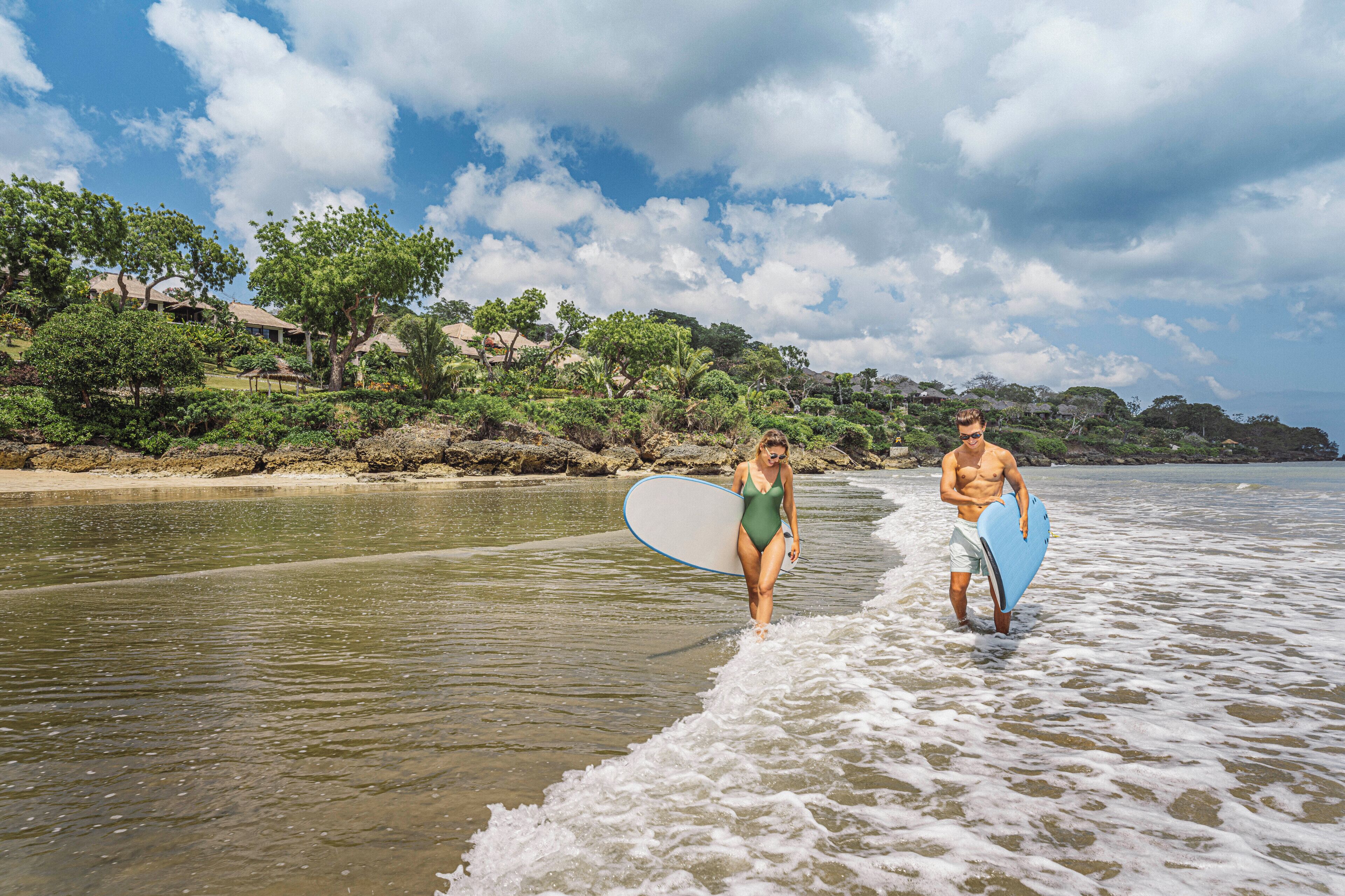 Duas pessoas entram na água com pranchas de surf, uma praia e um litoral rochoso com árvores ao fundo.