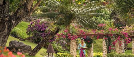 A woman in traditional attire standing under a stone archway with flowering plants.