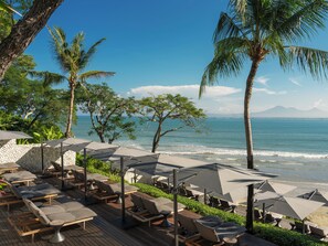 A beachside lounge area with sun loungers and umbrellas, overlooking the ocean and palm trees.