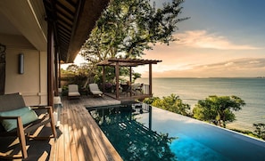 Una piscina infinita con terraza de madera, sillas de descanso y un pérgola con vista al mar.