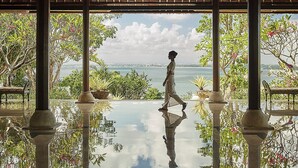 A person walking on a reflective floor under a wooden structure with columns, overlooking a body of water and greenery. - Four Seasons Resort Bali at Jimbaran Bay (Jimbaran)