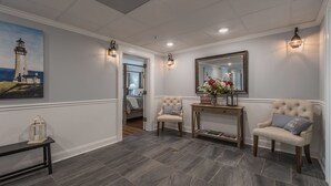 A hallway with a painting of a lighthouse, two upholstered chairs, a wooden console table with a vase of flowers, and a mirror on the wall.