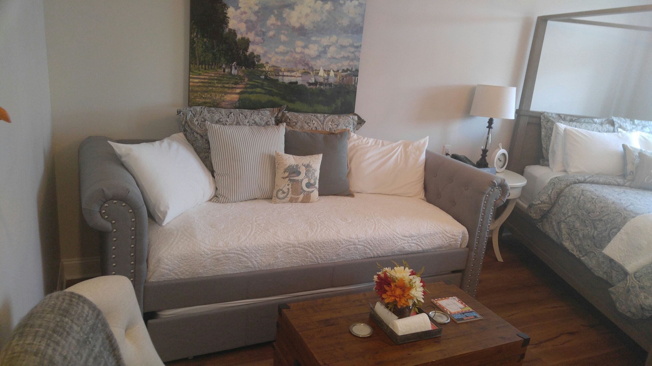 A cozy room with a grey sofa, a wooden coffee table with a vase of flowers, and a bed with a patterned blanket.