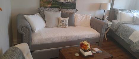 A cozy room with a grey sofa, a wooden coffee table with a vase of flowers, and a bed with a patterned blanket.