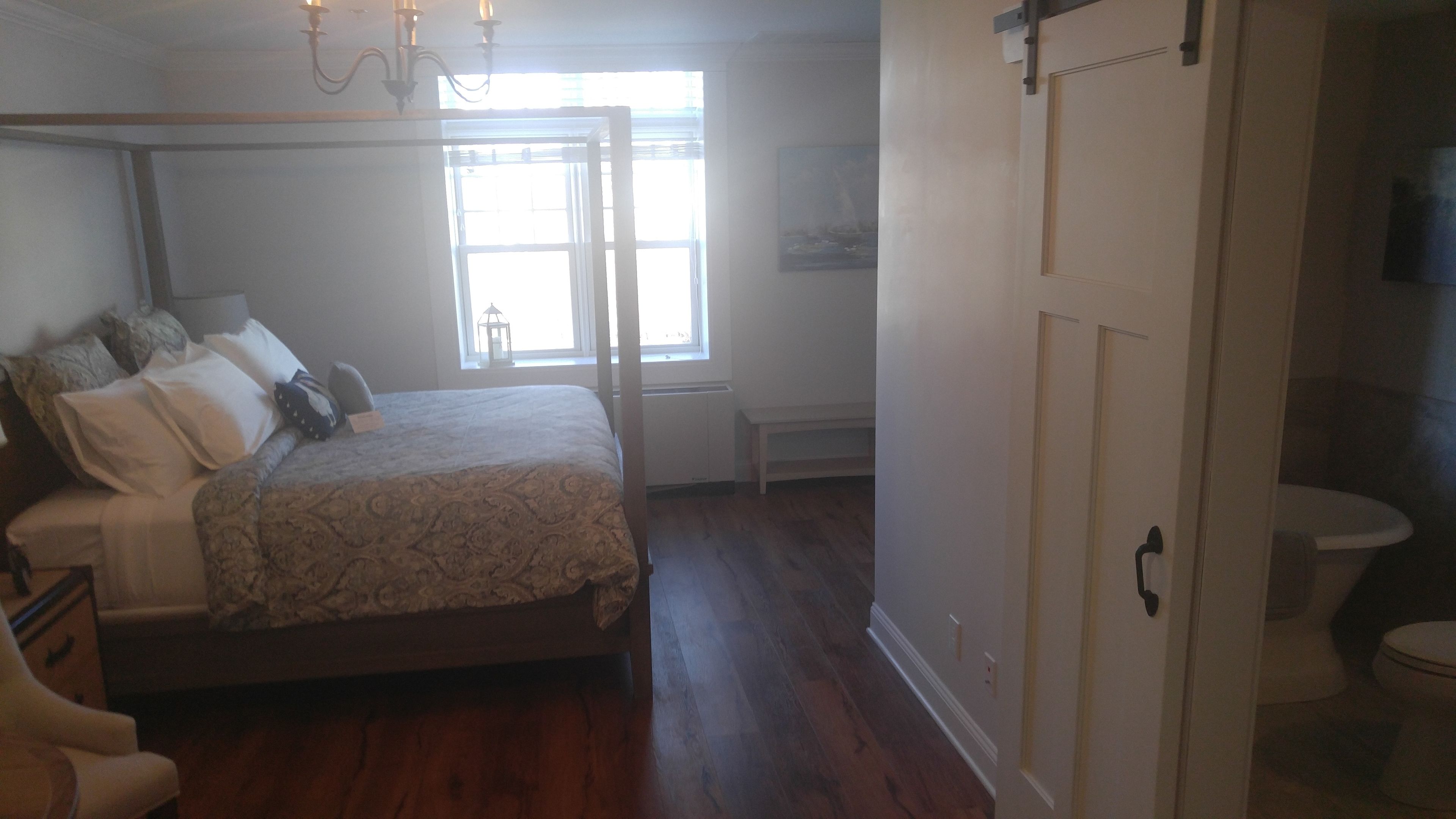 A bedroom with a bed, a nightstand, a chair, a window, and a door leading to a bathroom.