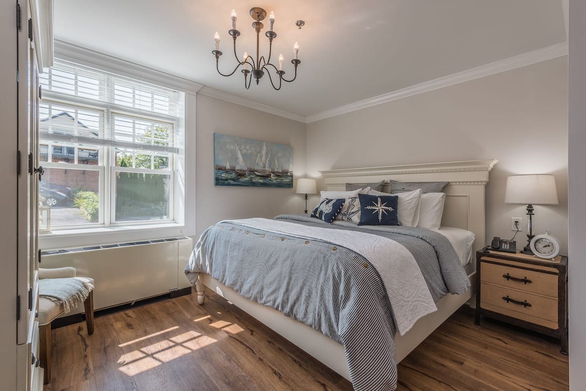 A bedroom with a large bed, a chandelier, a painting, a bedside table with a lamp, and a view of the outside through a window.