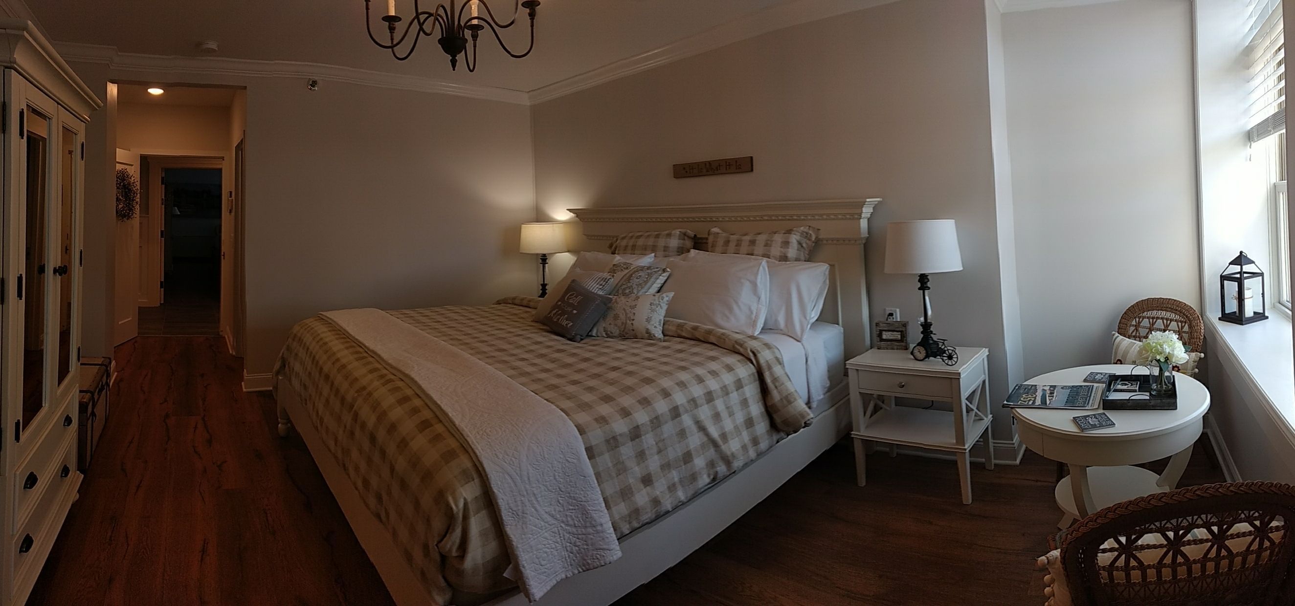 Un dormitorio con una cama grande, dos mesitas de noche con lámparas, una pequeña mesa redonda con un florero y una silla de mimbre.