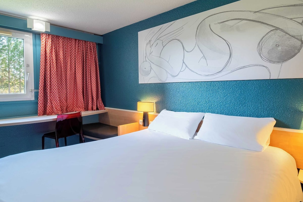 A hotel room with a bed, a desk, a chair, a lamp, and a large abstract painting on the wall.