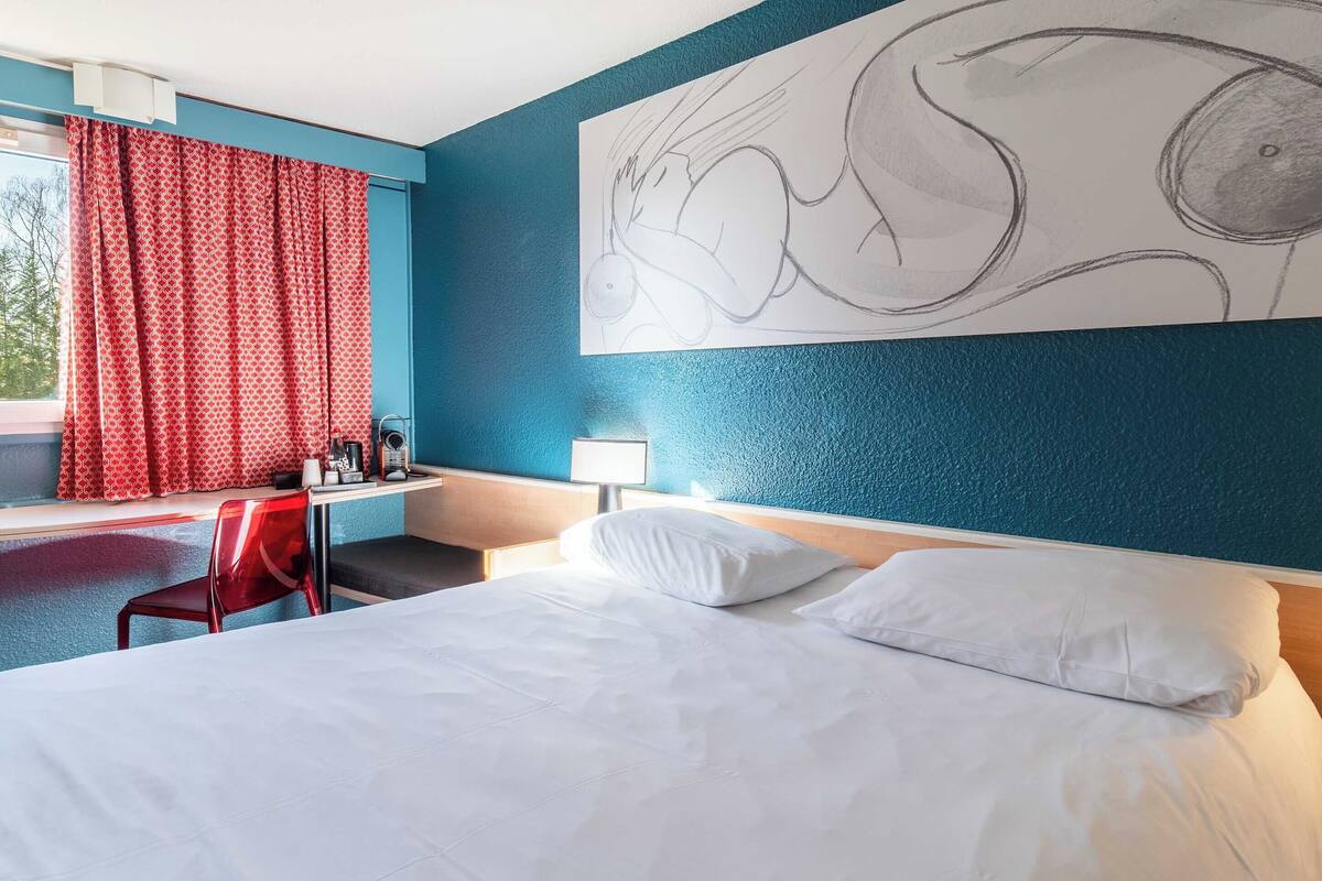 A hotel room with a bed, a desk with a chair, a window with red curtains, and a large abstract painting on the wall.