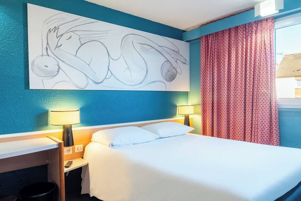 A hotel room with a bed, two bedside lamps, a wall art piece, and a window with red curtains.
