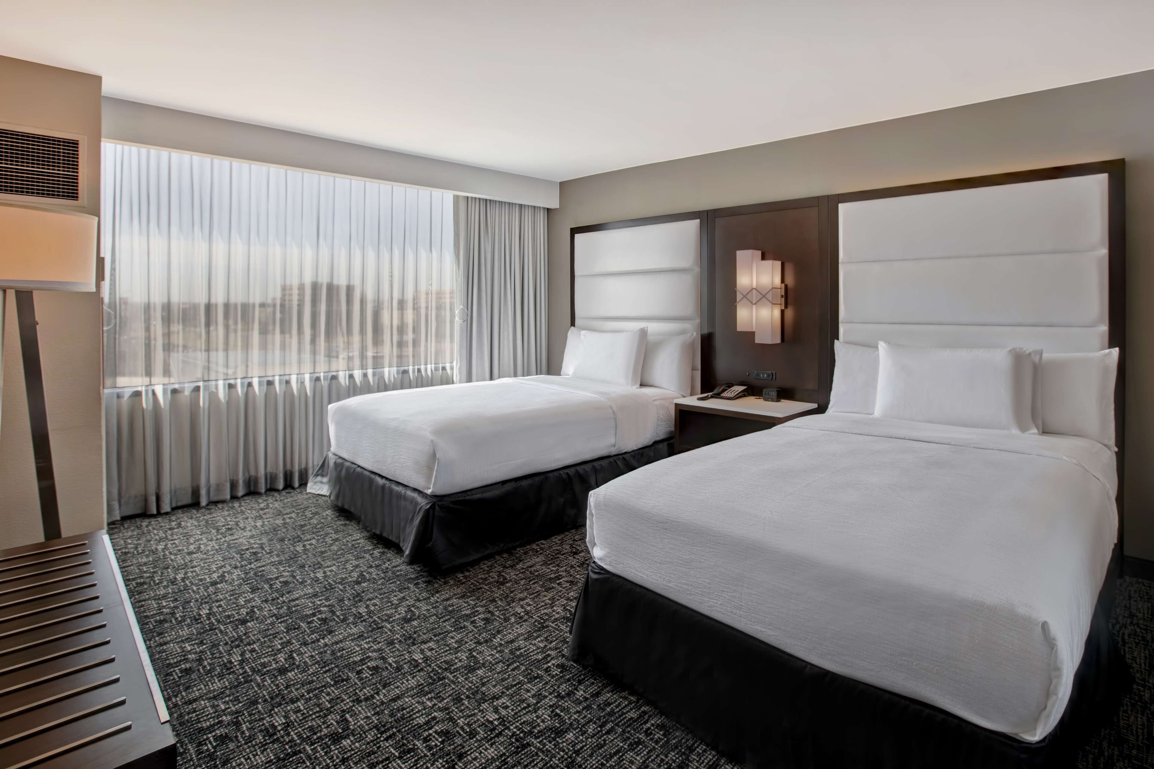 A hotel room with two beds, a large window with curtains, a bedside table with a lamp, and a wall-mounted air conditioning unit.