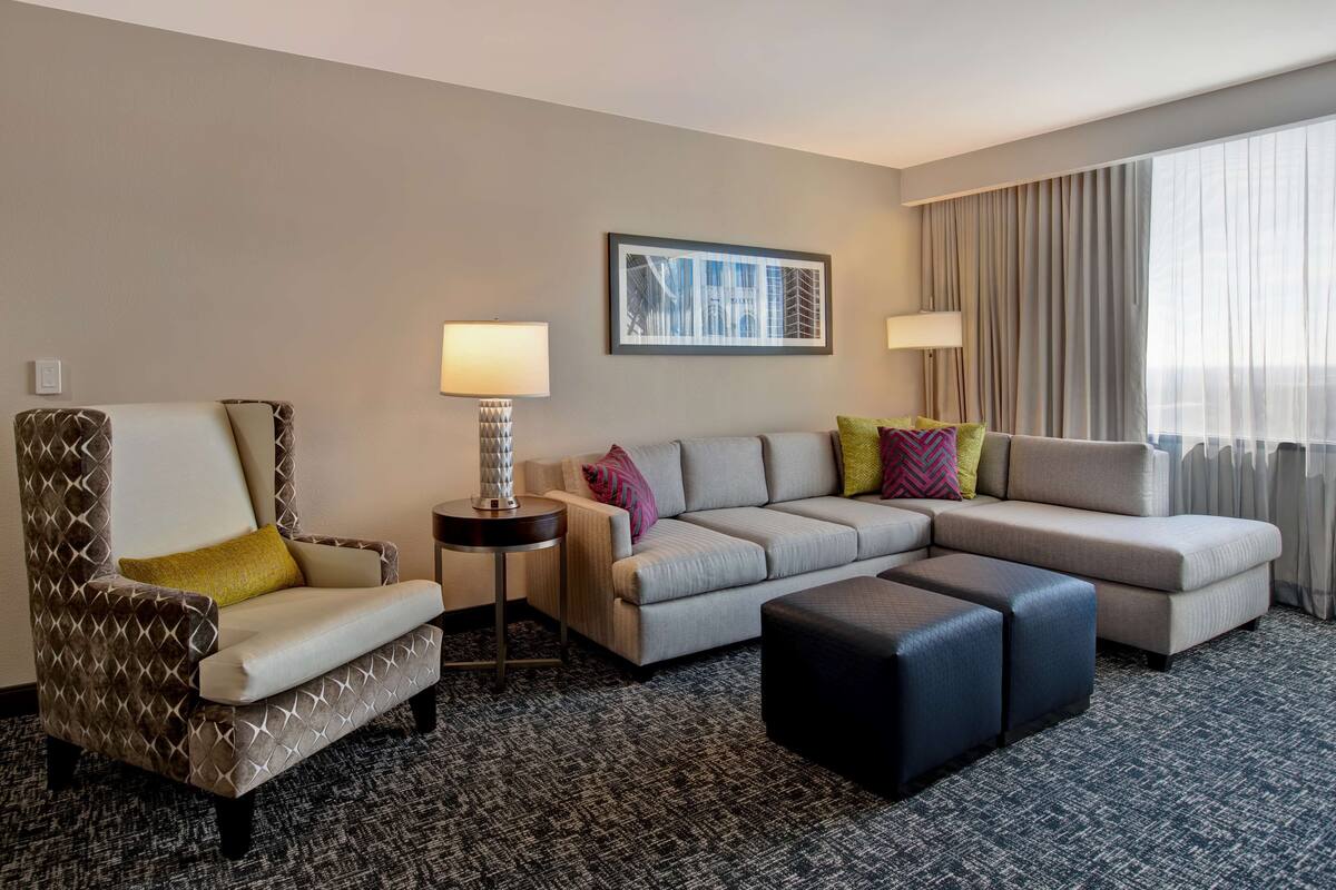 A hotel room with a patterned armchair, a beige sofa, a small side table with a lamp, and a large window with curtains.