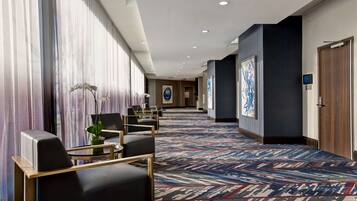 Un pasillo de hotel con alfombra estampada, zona de estar con sillas y una mesa, ventanas grandes con cortinas y obras de arte en las paredes.