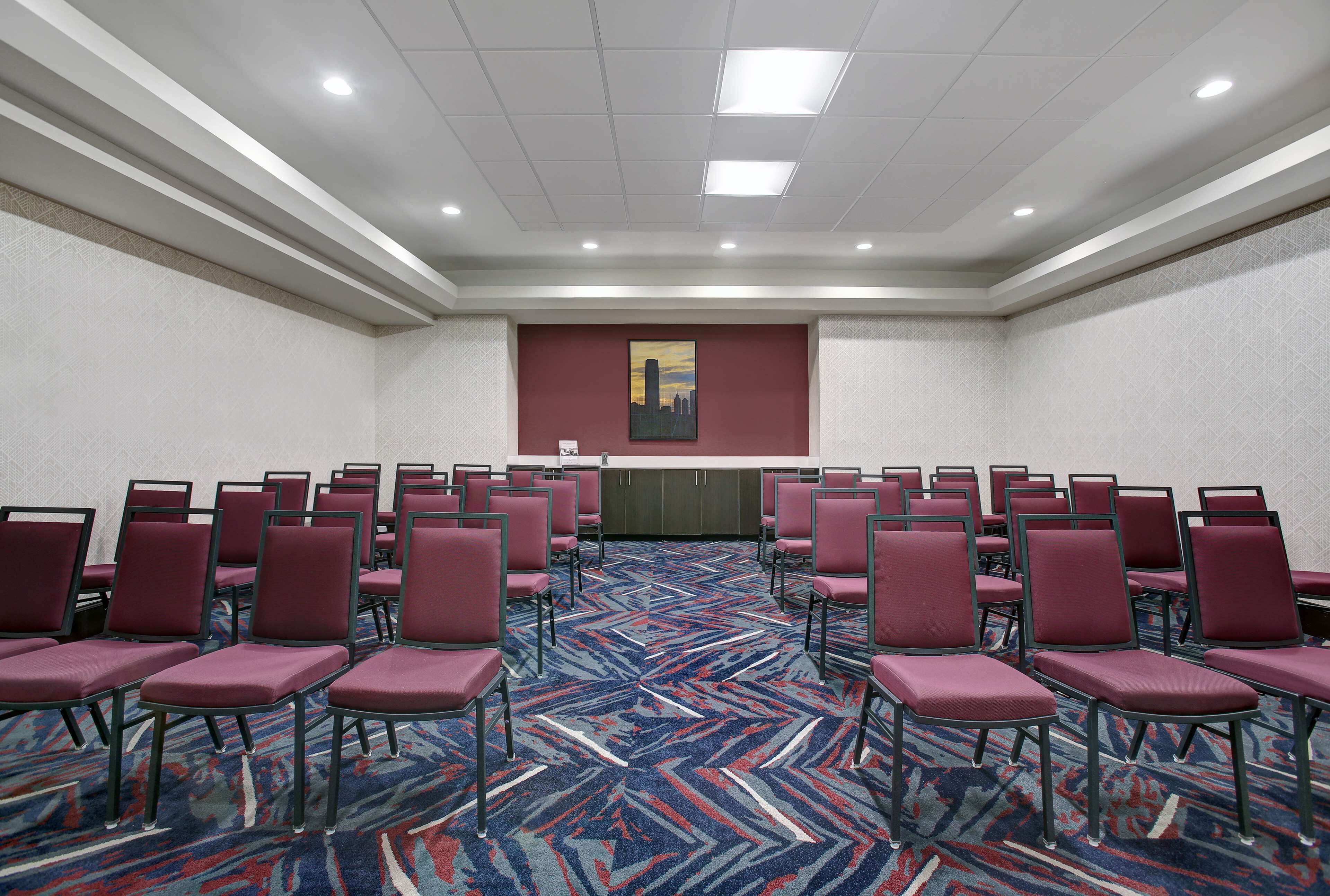 A conference room with rows of red chairs, a patterned carpet, and a cityscape painting on the wall.