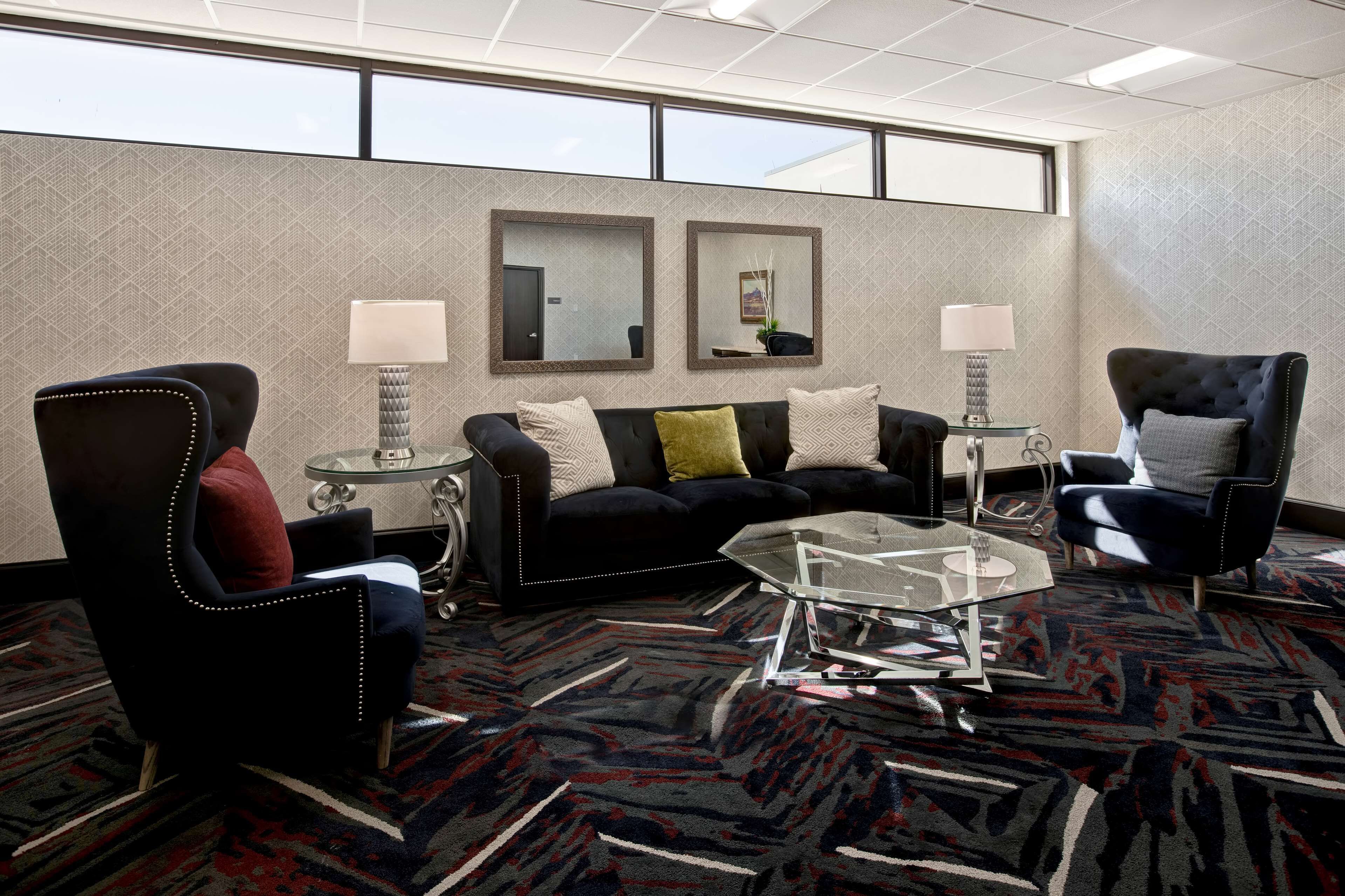 A modern hotel room with a black sofa, two armchairs, a glass coffee table, and two side tables with lamps. The room features a patterned carpet and large windows.