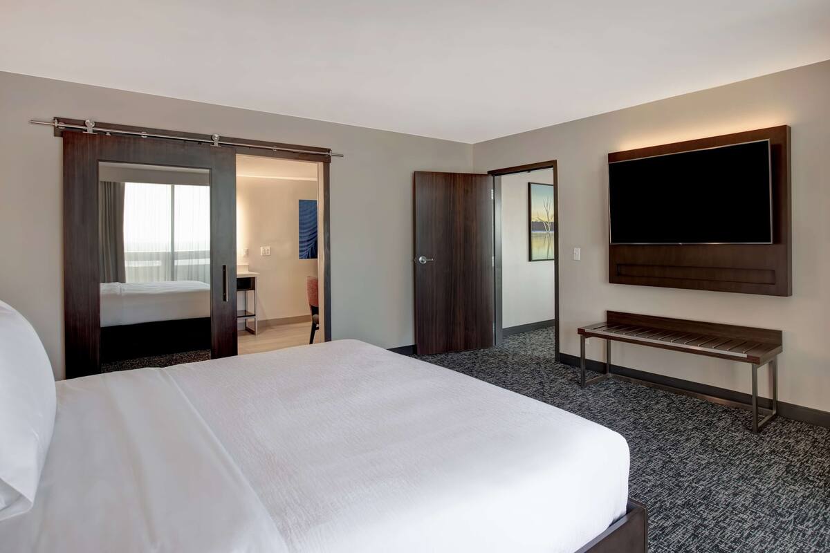 A hotel room with a large bed, a bench, a TV mounted on the wall, and a sliding door leading to a balcony.