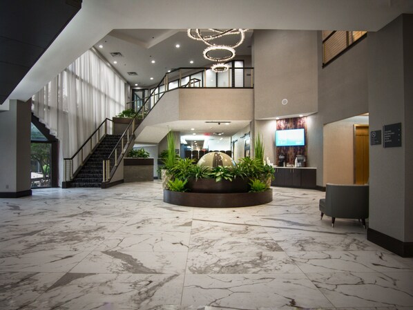 A spacious lobby with a marble floor, a central planter with greenery, a staircase, and a reception desk.
