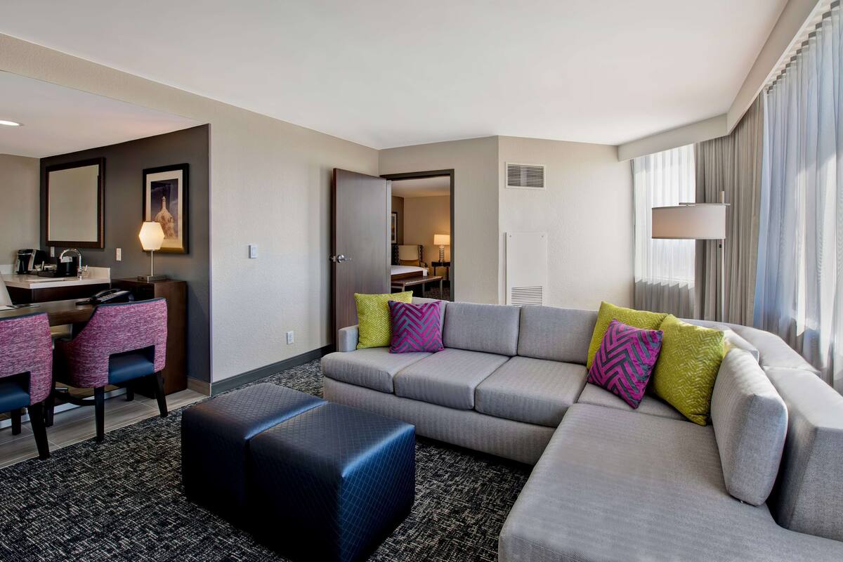 A modern hotel room with a grey sectional sofa, a small desk with a chair, a large mirror, and a window with curtains.