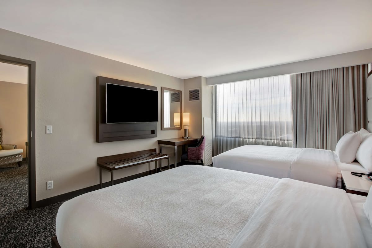 A hotel room with two beds, a desk, a TV, and a large window with curtains.