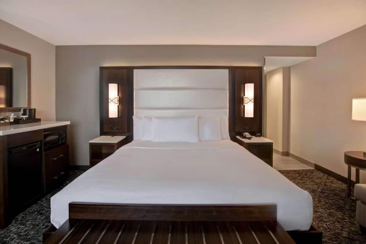 A hotel room with a large bed, a desk with a chair, a mirror, and a nightstand with a lamp.