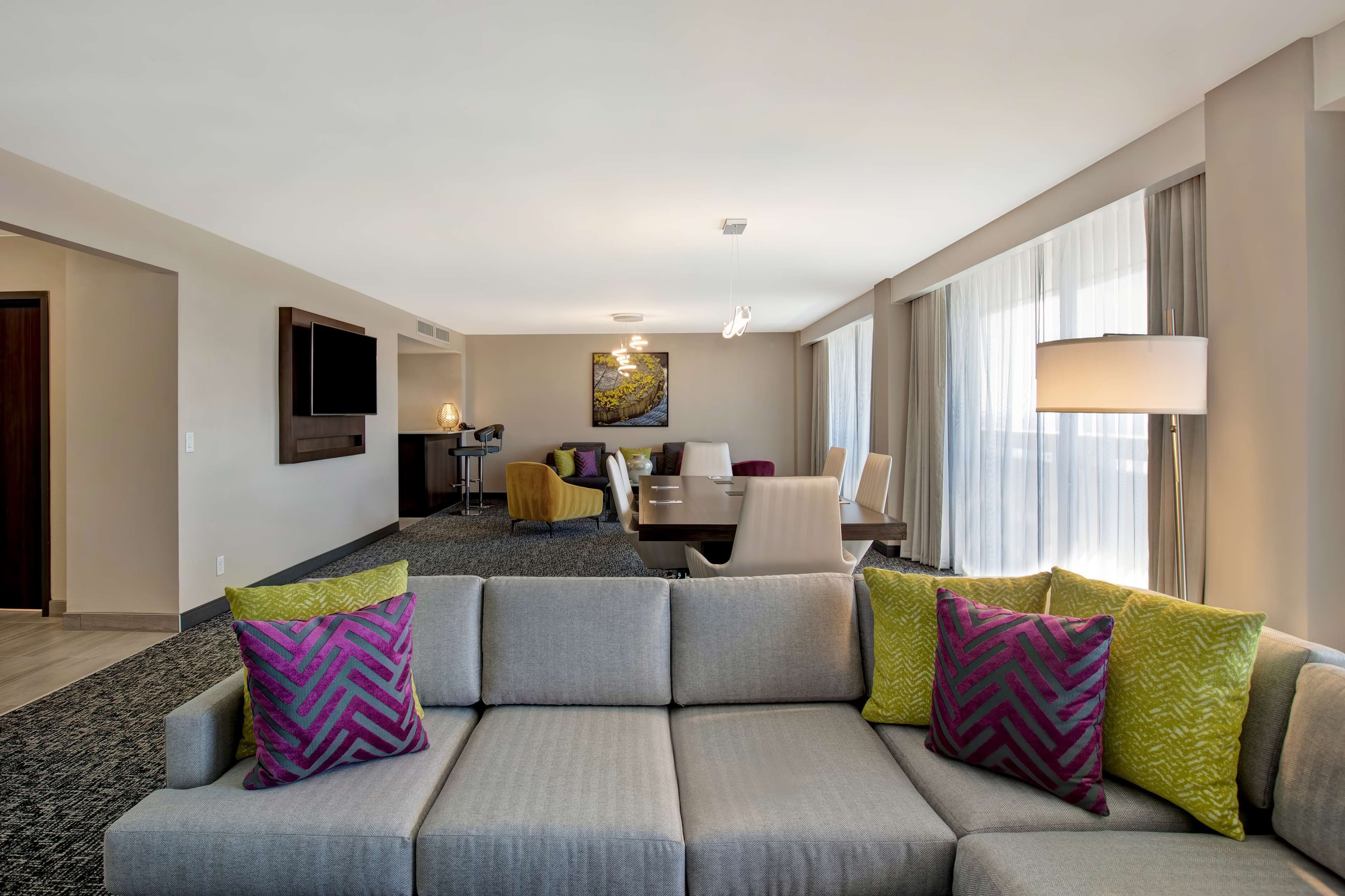A modern hotel room with a grey sofa, a dining area, and a large window with curtains.
