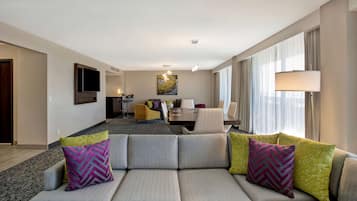 A modern hotel room with a grey sofa, a dining area, and a large window with curtains.