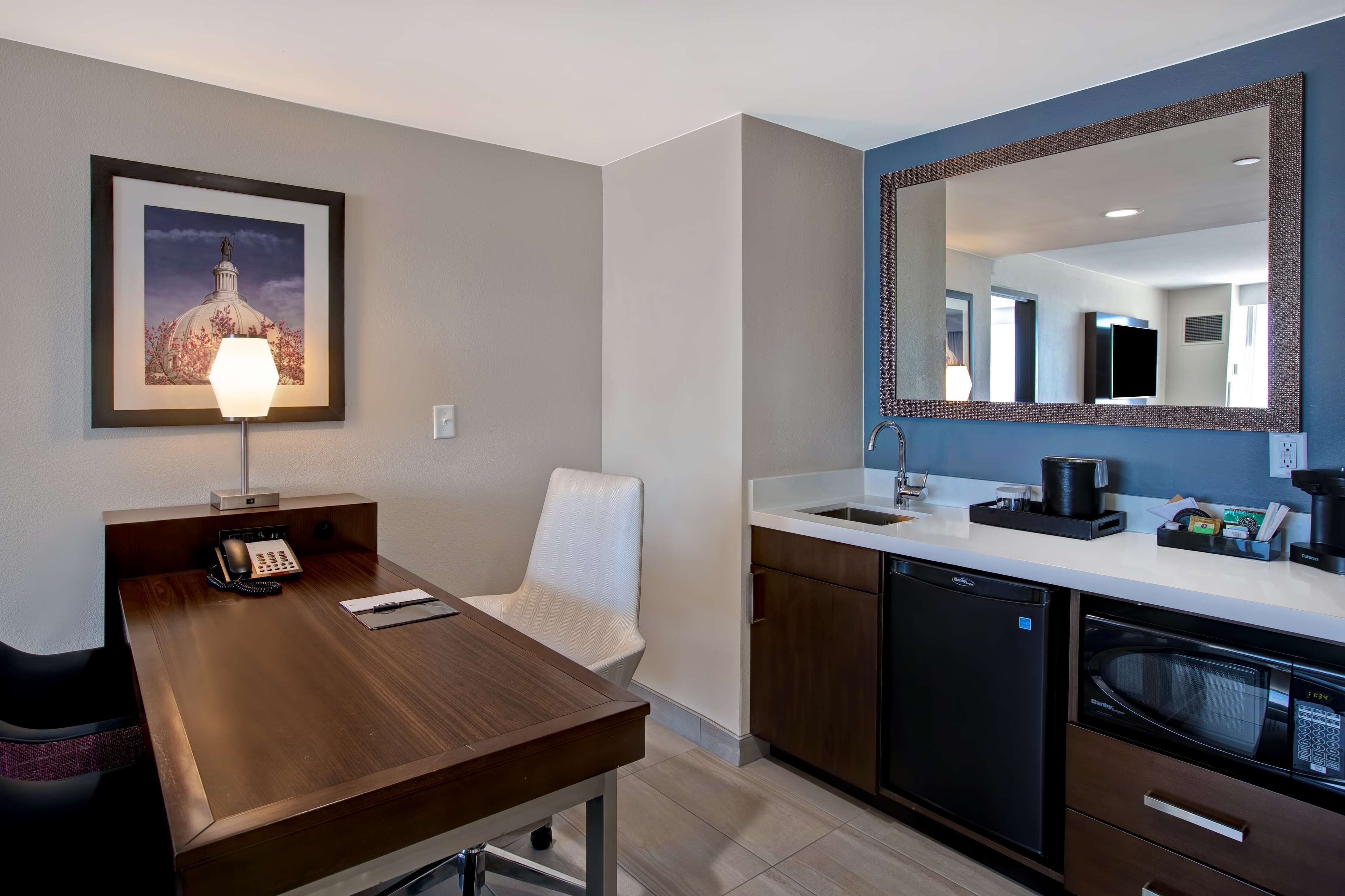 A modern hotel room with a wooden desk, a white chair, a large mirror, a kitchen area with a sink, a dishwasher, and a microwave.