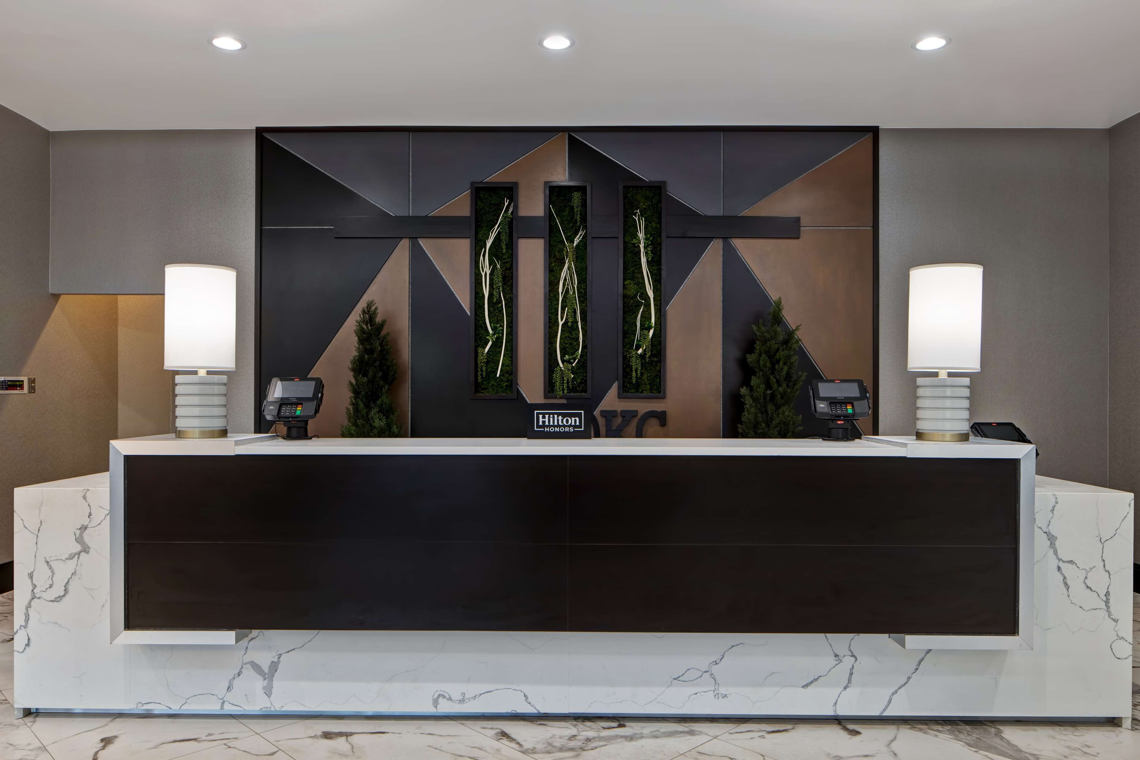 Un comptoir d’accueil moderne pour hôtel, doté d’un socle en marbre, de deux lampes murales, d’un terminal de paiement numérique et d’un mur décoratif orné de motifs abstraits et de plantes.